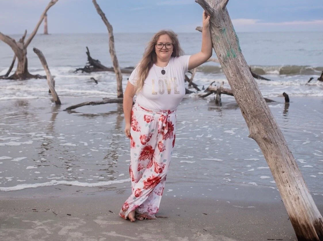 Woman standing on a beach holding a driftwood tree, with ocean waves and fallen trees in the background.